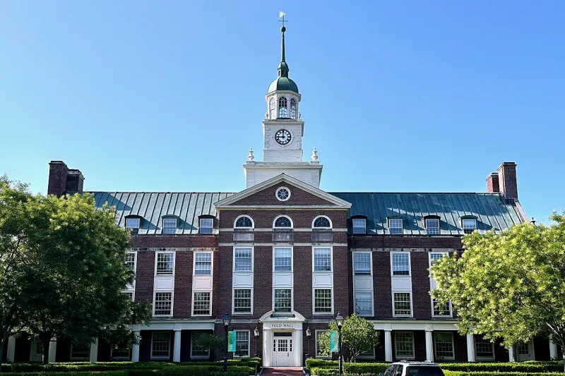 Langgestrecktes Backsteingebäude mit weißem Uhrturm unter blauem Himmel, umgeben von Bäumen